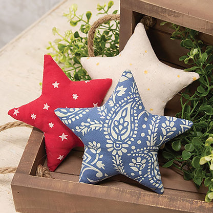 Americana plush star ornament set with red cream and blue patriotic fabric patterns displayed in rustic wooden tray with greenery