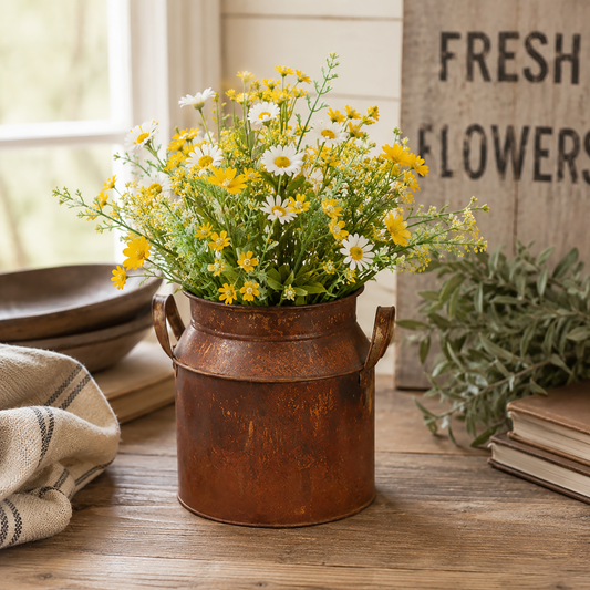 Rusty tin wide milk can filled with yellow and white wildflowers displayed on a rustic farmhouse tabletop with greenery and vintage décor accents.