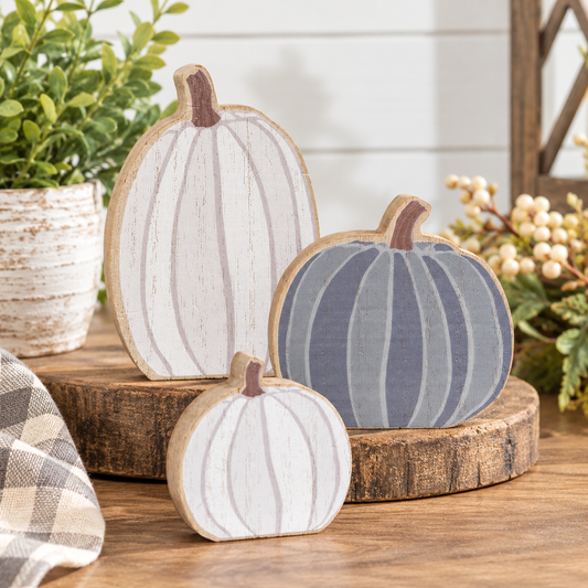 Three rustic wooden pumpkin decorations in white, gray, and slate blue, styled on a wooden surface with a potted plant and autumn decor in the background.