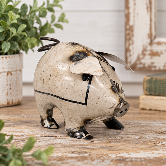 Small handcrafted white recycled metal pig figurine with distressed black markings, exposed metal accents, dimensional ears, black eyes, round snout, curled metal tail, and dark metal feet, displayed on a weathered wood tabletop with soft greenery, vintage books, and neutral farmhouse-style decor in the background.