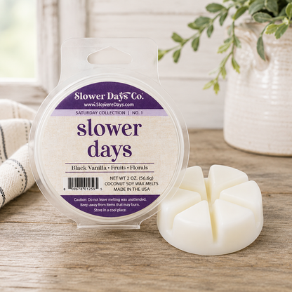 A round plastic tub of Slower Days Co. soy wax melts with a clear hanger window and a white multi-wool melt stick placed beside it on a wooden surface.