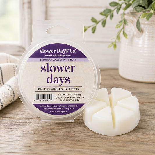 A round plastic tub of Slower Days Co. soy wax melts with a clear hanger window and a white multi-wool melt stick placed beside it on a wooden surface.