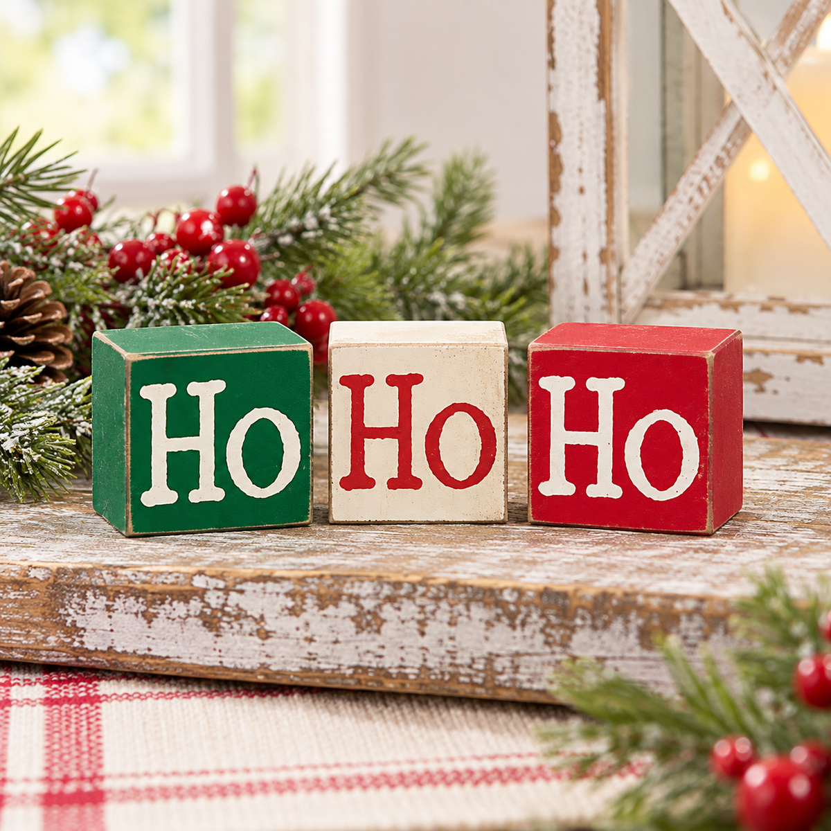 Three wooden decorative blocks spelling out 'Ho Ho Ho' in white lettering, with one green block, one cream block, and one red block, styled on a rustic wooden surface with pine branches and red berries.