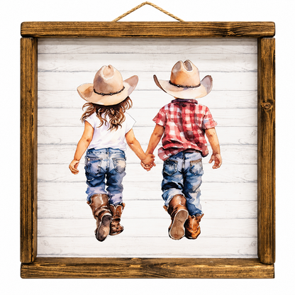 A square framed art print featuring a watercolor-style illustration of two children in cowboy hats and jeans holding hands, viewed from behind, set against a white wood plank background. The artwork is enclosed in a rustic brown wooden frame with a hanging rope at the top.