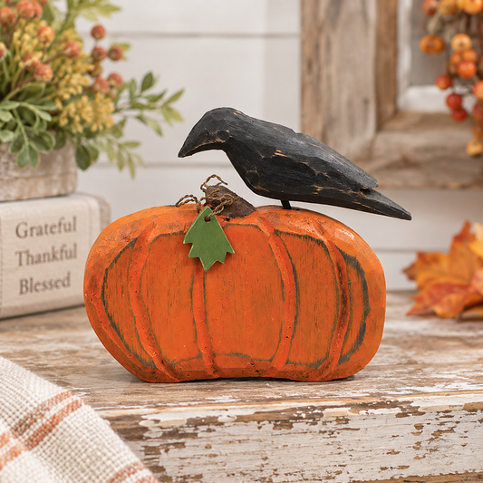 A decorative orange pumpkin figurine with a distressed finish, topped with a black bird sculpture and a small green leaf accent, displayed on a rustic wooden surface with fall decor in the background.