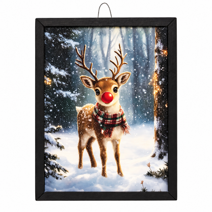 Framed wall art featuring a snowy winter forest scene with a brown reindeer wearing a red nose and plaid scarf, displayed in a black frame against a white background.