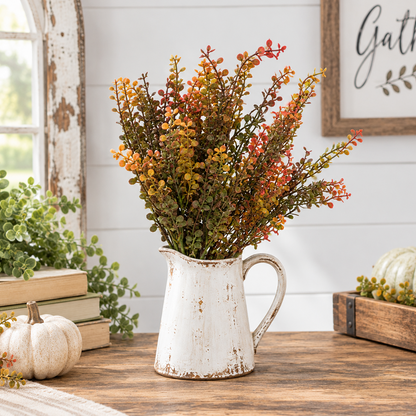 A rustic white distressed ceramic pitcher vase filled with autumn artificial foliage in orange, yellow, and green, displayed on a wooden table in a styled home decor setting.