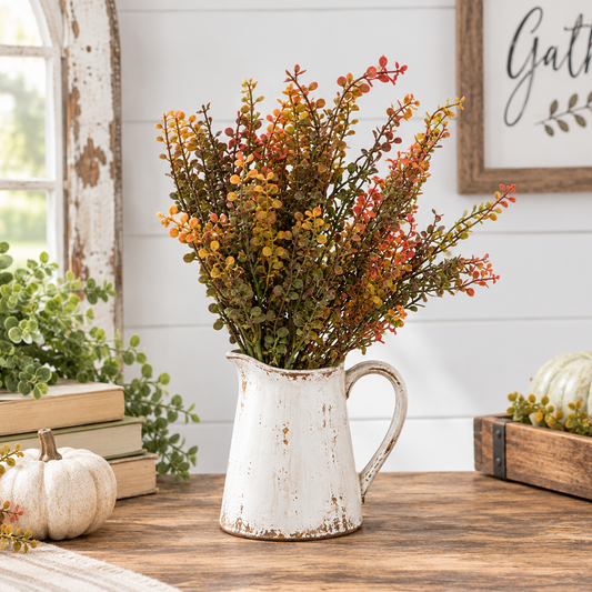 A rustic white distressed ceramic pitcher vase filled with autumn artificial foliage in orange, yellow, and green, displayed on a wooden table in a styled home decor setting.
