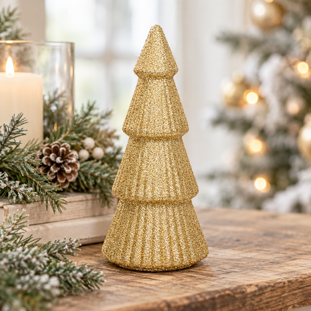 A small tiered Christmas tree decoration in textured gold glitter sits on a wooden table, surrounded by holiday greenery, a lit candle, and a softly blurred decorated Christmas tree in the background.