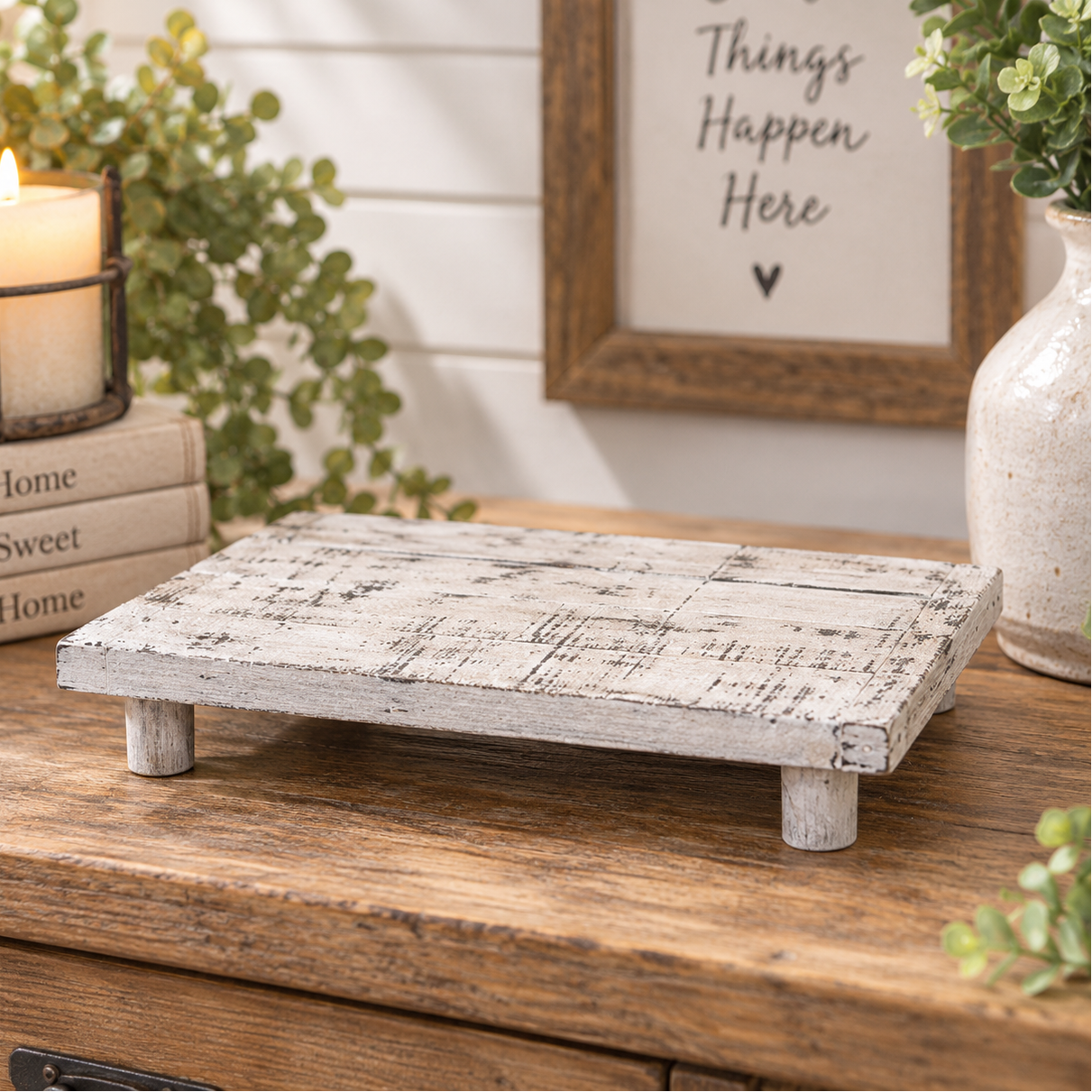 A rectangular wooden stand or tray with a distressed white and gray rustic finish, shown on a wooden table in a styled home decor setting with a candle, books, plants, and framed wall art.