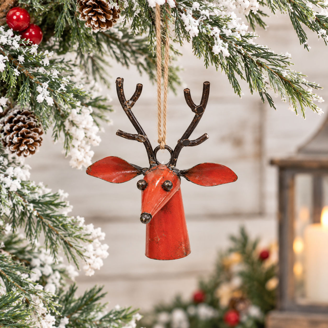 A red metal reindeer bell ornament with dark brown antlers hangs from a tree branch decorated with pinecones, white snow-like faux snow, and red berries, with a softly blurred holiday background.