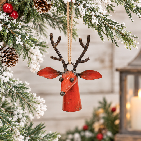 A red metal reindeer bell ornament with dark brown antlers hangs from a tree branch decorated with pinecones, white snow-like faux snow, and red berries, with a softly blurred holiday background.