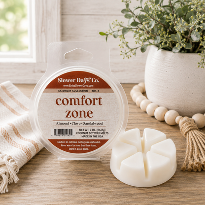 A packaged round wax melt container labeled 'Slower Days Co. comfort zone' stands on a wooden surface beside a white wax melt piece.