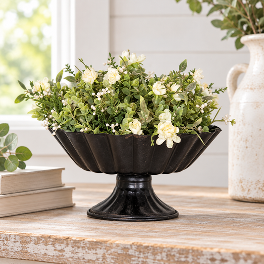 Black scalloped metal pedestal urn filled with cream flowers and greenery on a rustic wood table in bright farmhouse-style decor.