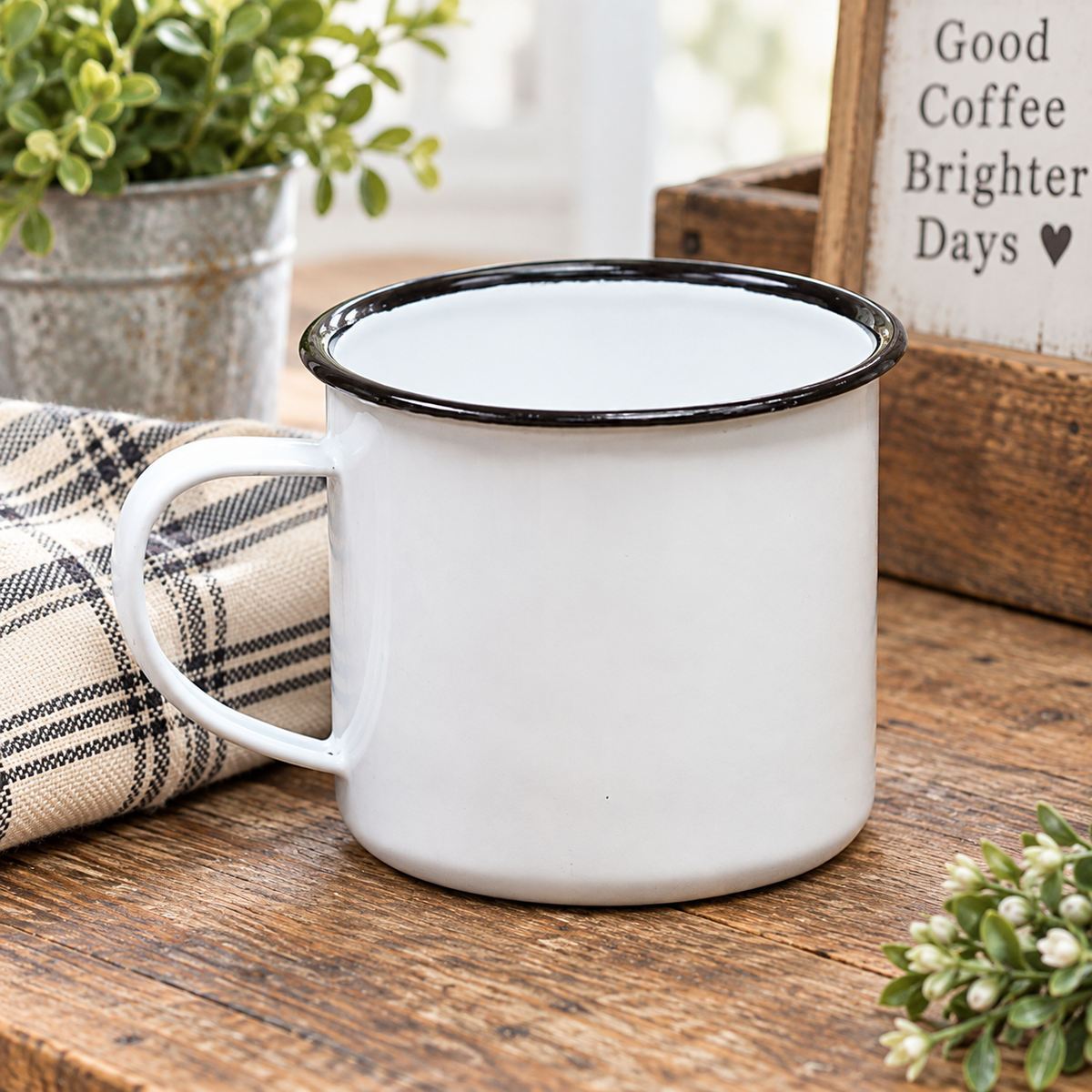 A white enamel-style mug with a black rim sits on a rustic wooden table, styled with a plaid napkin, a potted plant, and a wooden sign that reads 'Good Coffee Brighter Days'.