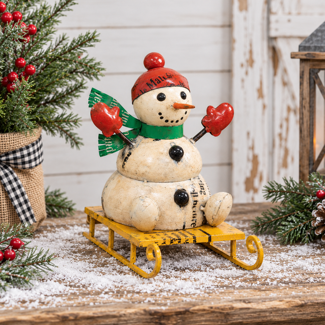 A decorative stacked-snowman figurine wearing a red hat and green scarf, sitting on a yellow sled, styled on a rustic wooden surface with holiday greenery, faux snow, pinecones, and a lantern in the background.