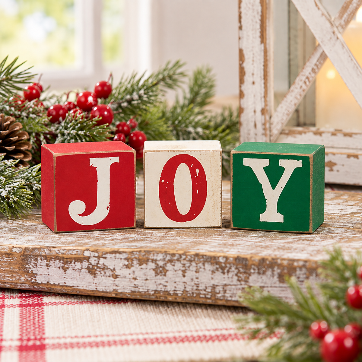 Three wooden decorative blocks spelling JOY in red, cream, and green with white lettering, styled on a rustic wooden surface with evergreen branches and red berries.