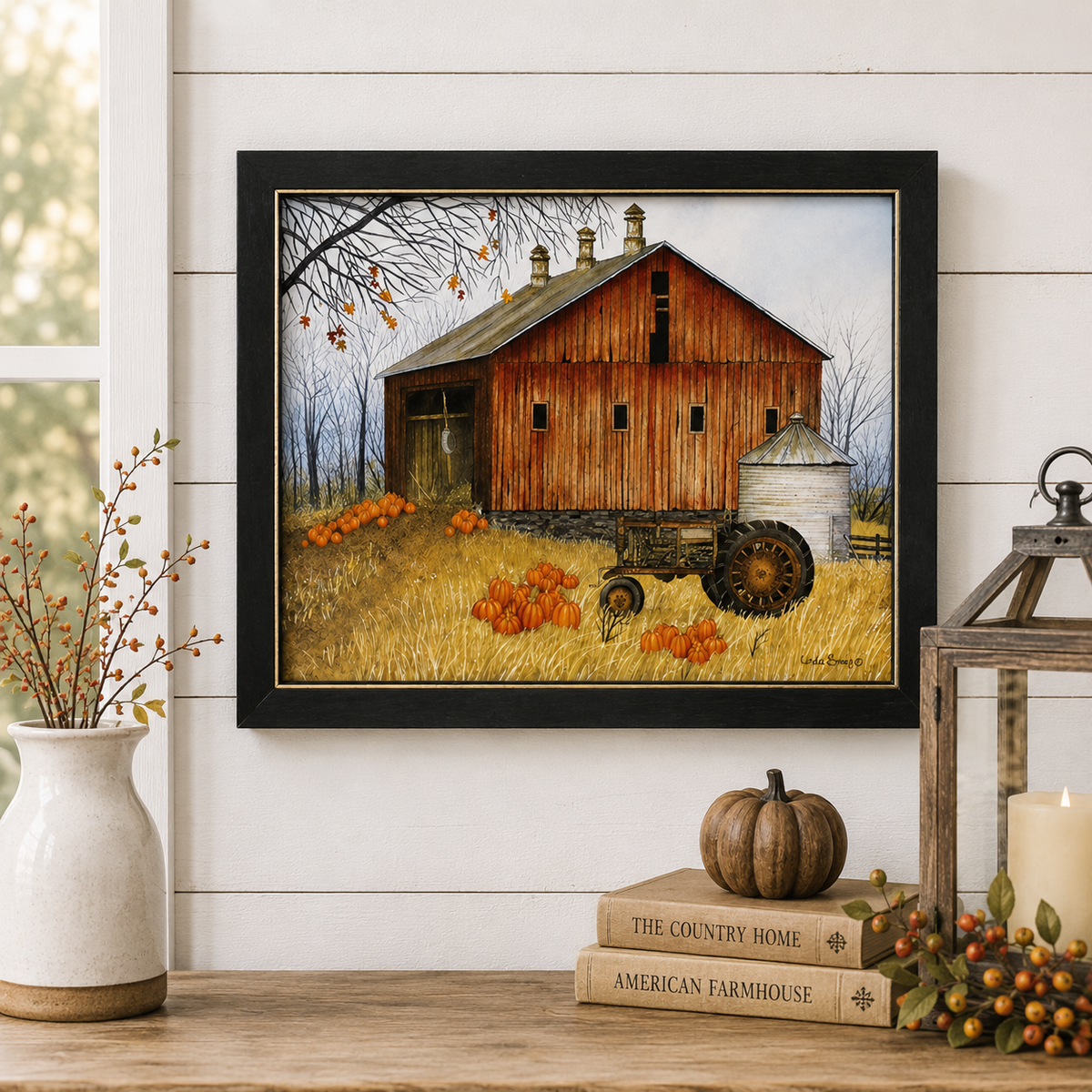 Framed wall art showing a rustic red barn, silo, and pumpkins in a fall field, displayed on a white shiplap wall above a wooden surface with a vase, books, a pumpkin, and a lantern.