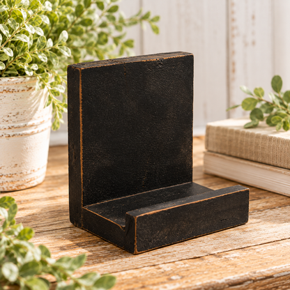 A rustic black wooden L-shaped bookend or desk stand with distressed edges, shown on a wooden tabletop with potted plants and books in the background.
