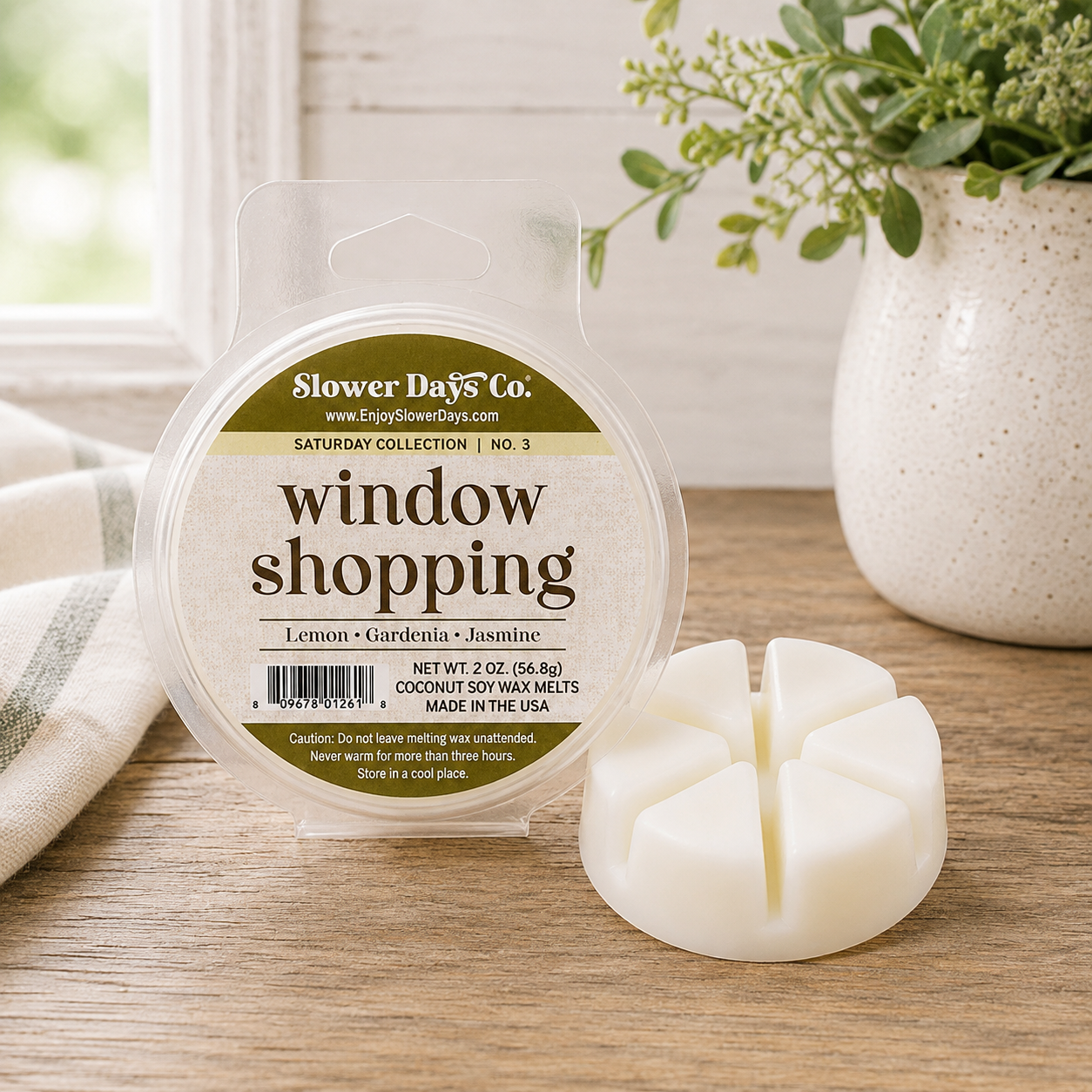 A packaged coconut soy wax melt from Slower Days Co. labeled 'window shopping' (Lemon & Jasmine), shown next to its three separate wax melts on a wooden tabletop with a vase of greenery in the background.