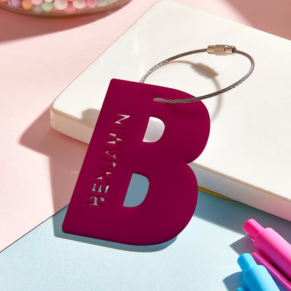 A close-up view of a 3D printed luggage tag shaped like the letter "B" in a deep wine red color, highlighting its smooth acrylic finish and sturdy design for easy identification.