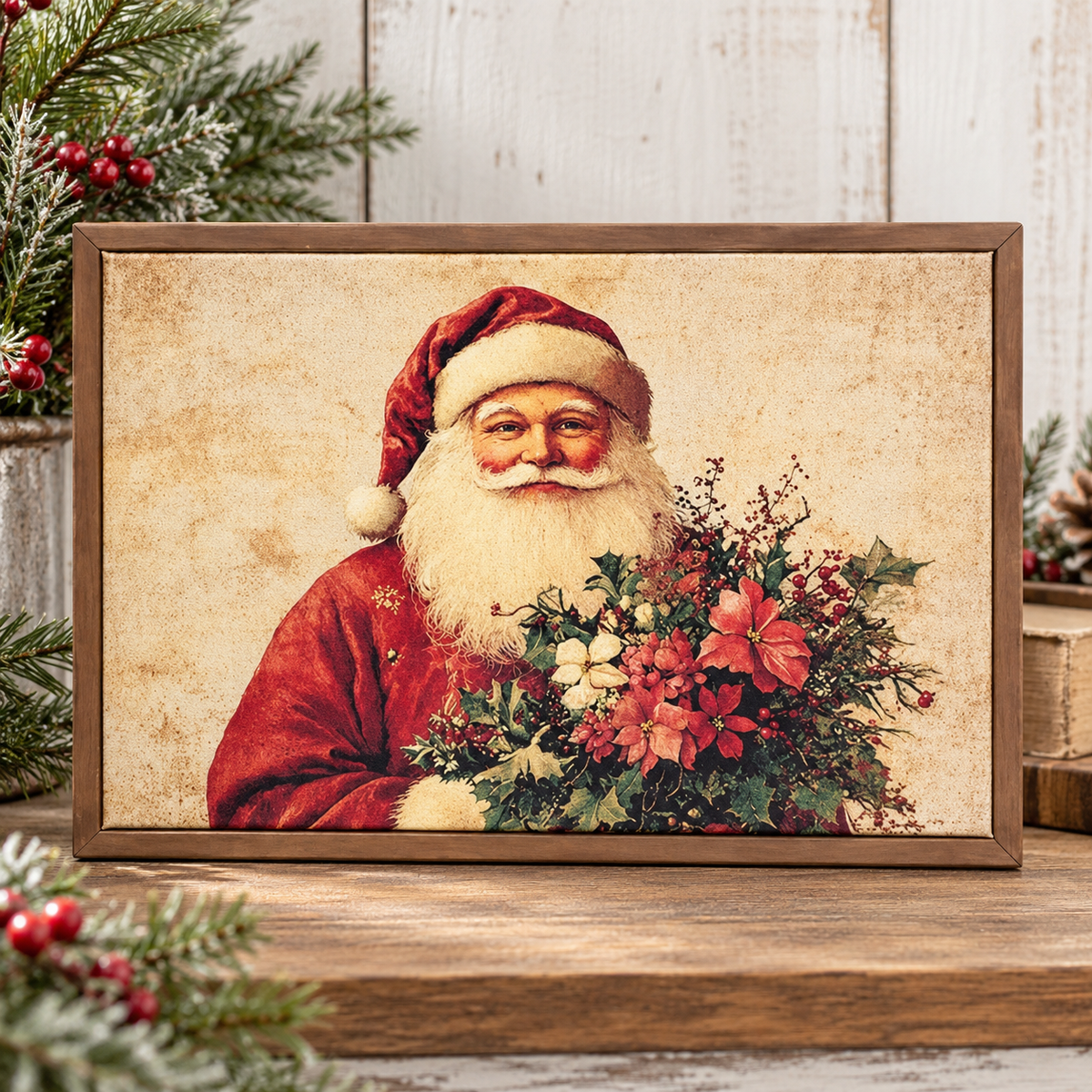 A framed vintage-style Christmas print showing Santa Claus in a red suit and hat holding a bouquet of red and white flowers, displayed on a wooden surface with holiday greenery and rustic decor around it.