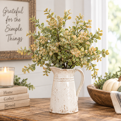 An artificial variegated green and yellow leafy plant arrangement in a distressed white ceramic pitcher, styled on a wooden table with a candle, books, and a bowl of pumpkins.