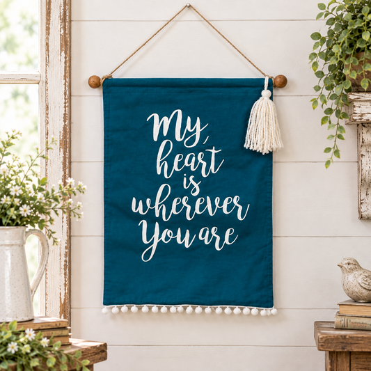 A teal fabric wall hanging with white script text reading 'My heart is wherever You are,' suspended from a wooden rod with a cream tassel and pom-pom trim, displayed against a white shiplap wall with rustic decor.
