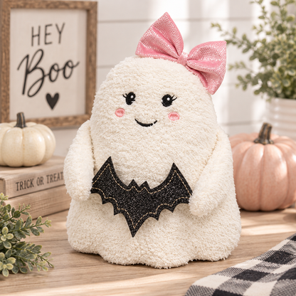 A plush Halloween ghost decoration in cream sherpa fabric with a pink glitter bow on its head, a black glitter bat-wing motif on the front, and embroidered facial features, displayed on a wooden surface with pumpkins and fall decor in the background.