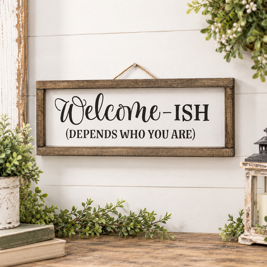 A rustic wooden wall sign with the phrase 'Welcome-ISH (DEPENDS WHO YOU ARE)' in black script and serif lettering, hanging on a white shiplap wall above a wooden surface with greenery and decor.