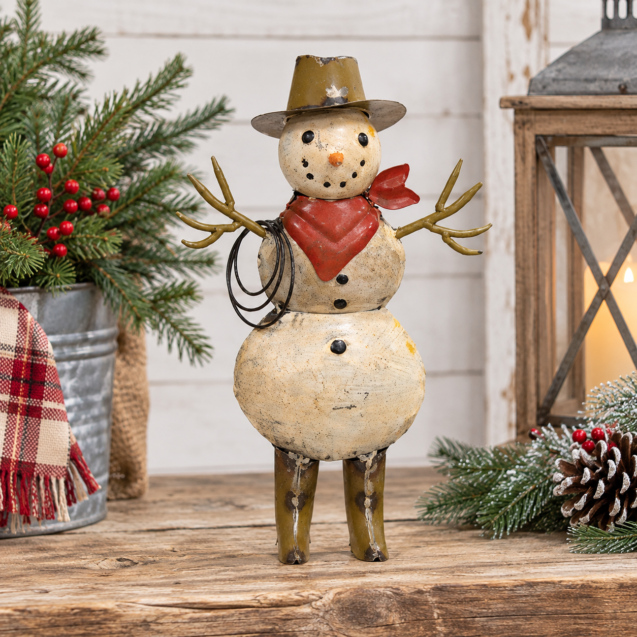 A rustic painted snowman figurine standing on a wooden surface, wearing a brown hat, red bandana, and holding a lasso. The scene is styled with evergreen branches, red berries, a lantern, and a plaid throw blanket in the background.