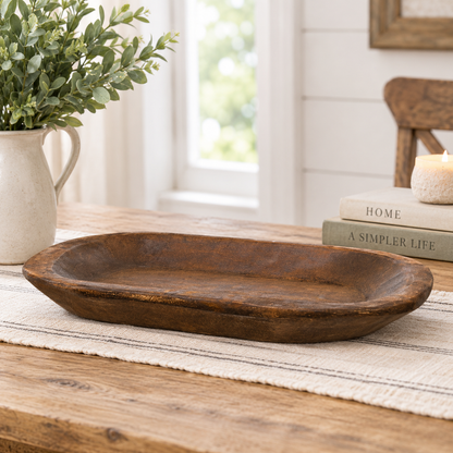 A rustic oval wooden serving tray with a distressed dark brown finish, displayed on a woven runner on a wooden table in a bright, styled room setting.