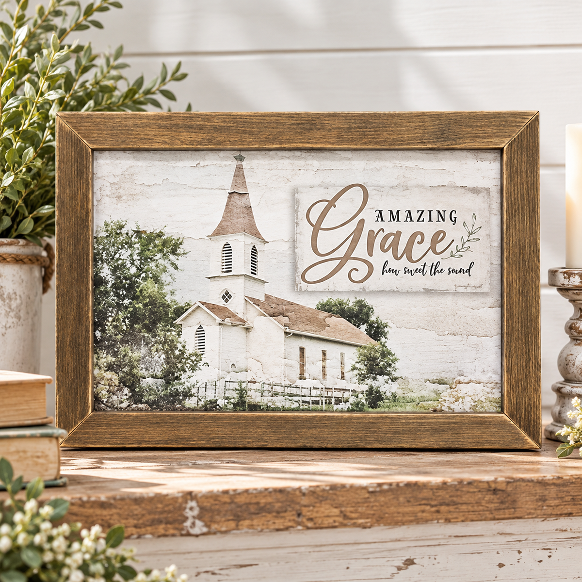 A framed decorative print with a rustic brown wood frame sits on a wooden surface. The print shows a white church with trees and a sign that reads “Amazing Grace how sweet the sound.