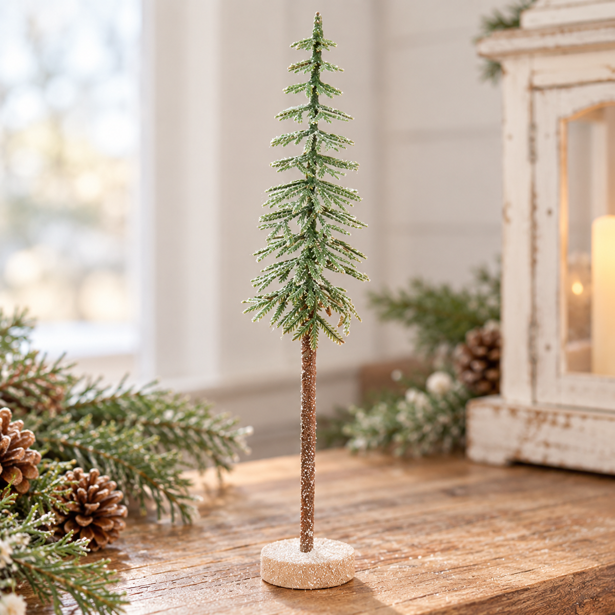 A small flocked green Christmas tree with a brown textured stem and round glittery base, displayed on a wooden table with pinecones and holiday greenery in a softly lit indoor setting.