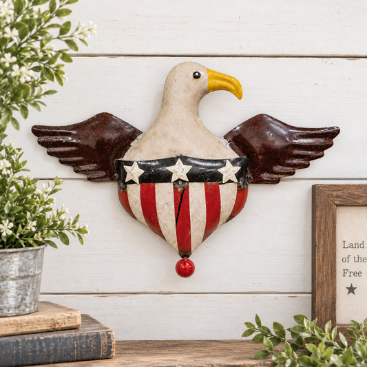 A decorative metal wall hanging shaped like an eagle with spread wings and a red, white, and blue striped heart-shaped body, displayed against a white wood plank wall with plants and a framed print nearby.