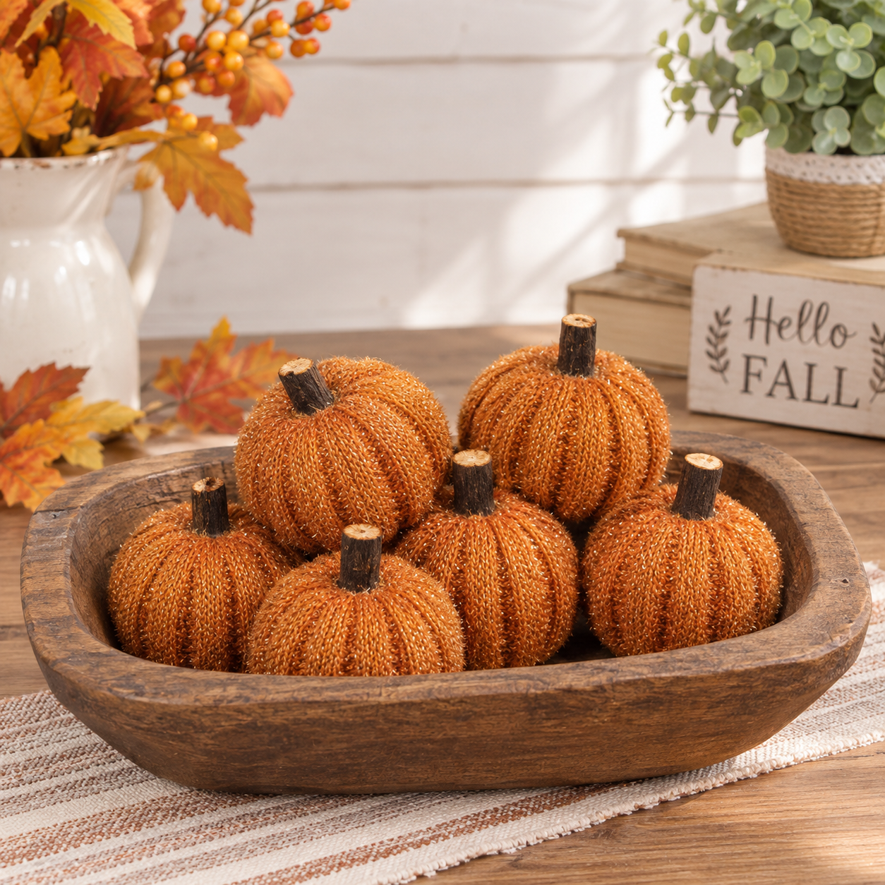Six decorative faux pumpkins in a rustic wooden bowl, styled on a table with autumn leaves, a white pitcher, books, and a small plant in the background.