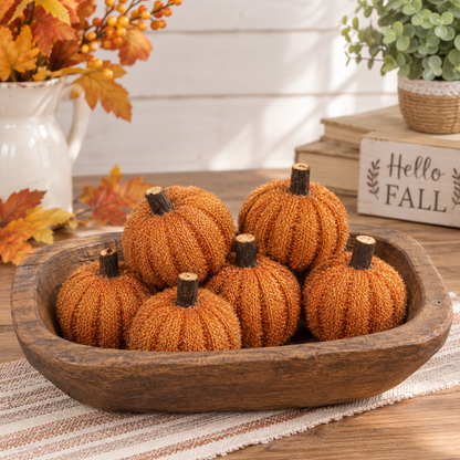 Six decorative faux pumpkins in a rustic wooden bowl, styled on a table with autumn leaves, a white pitcher, books, and a small plant in the background.