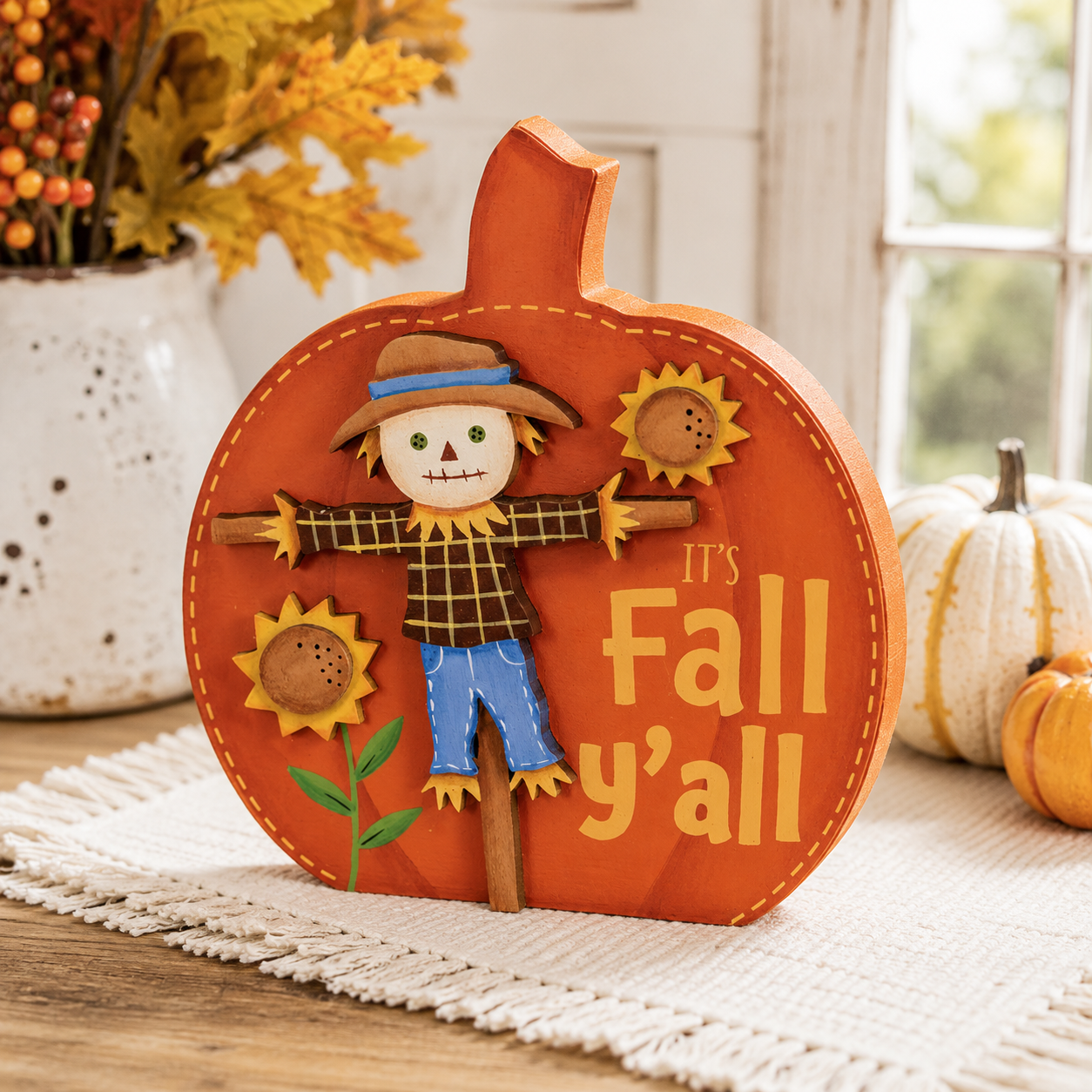 An autumn-themed wooden decorative sign shaped like a pumpkin, featuring a scarecrow, sunflowers, and the text 'It's Fall y'all' in a warm orange-brown tone, styled on a table runner with pumpkins and a vase of fall foliage in the background.