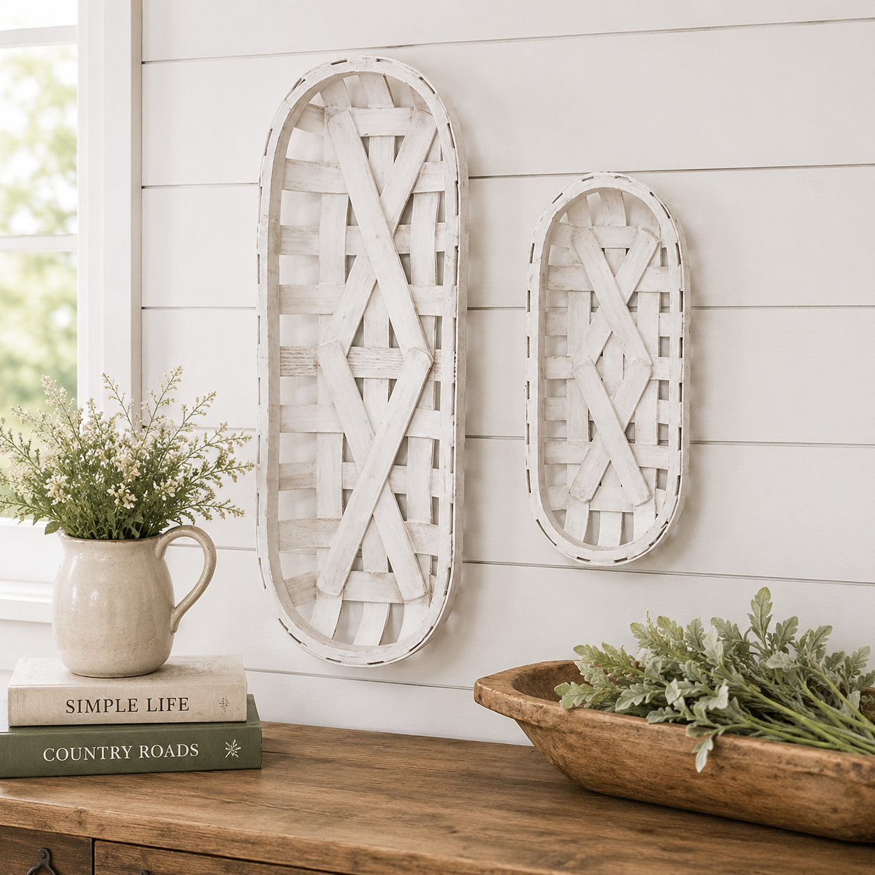Two white distressed wooden basket wall hanging decor pieces displayed on a white shiplap wall above a wooden console table with decorative books, a ceramic pitcher of flowers, and a wooden bowl.