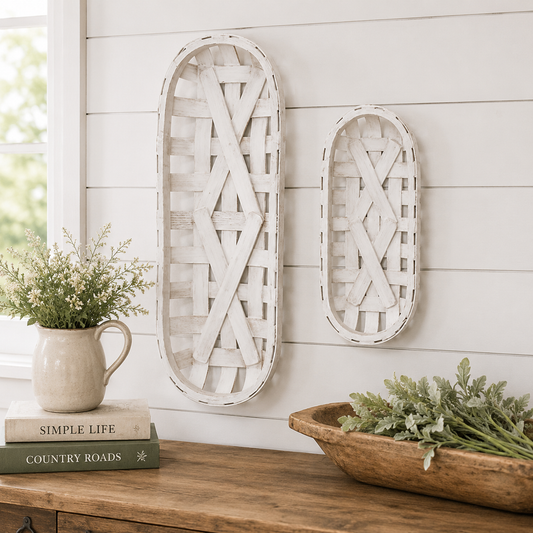 Two white distressed wooden basket wall hanging decor pieces displayed on a white shiplap wall above a wooden console table with decorative books, a ceramic pitcher of flowers, and a wooden bowl.