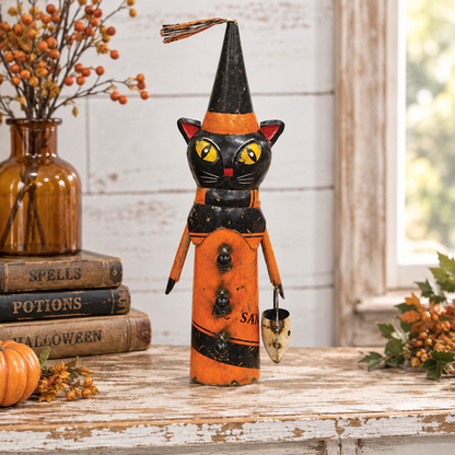 A decorative black and orange cat-shaped Halloween ornament with yellow eyes stands on a rustic wooden table. The figure wears a pointed black hat with orange bands, holds a small metal bell, and is styled with autumn props including books, a vase, berries, and a window in the background.