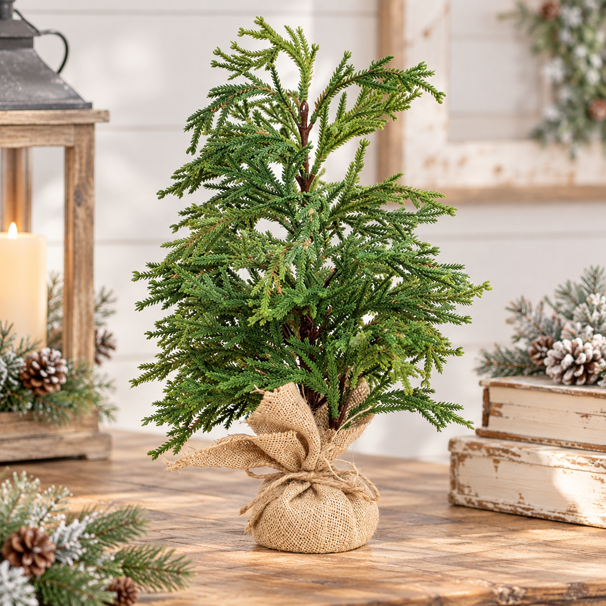 A small artificial evergreen tree in a burlap-wrapped pot sits on a wooden table, styled with holiday greenery, a lantern, candles, and stacked books in a rustic indoor setting.