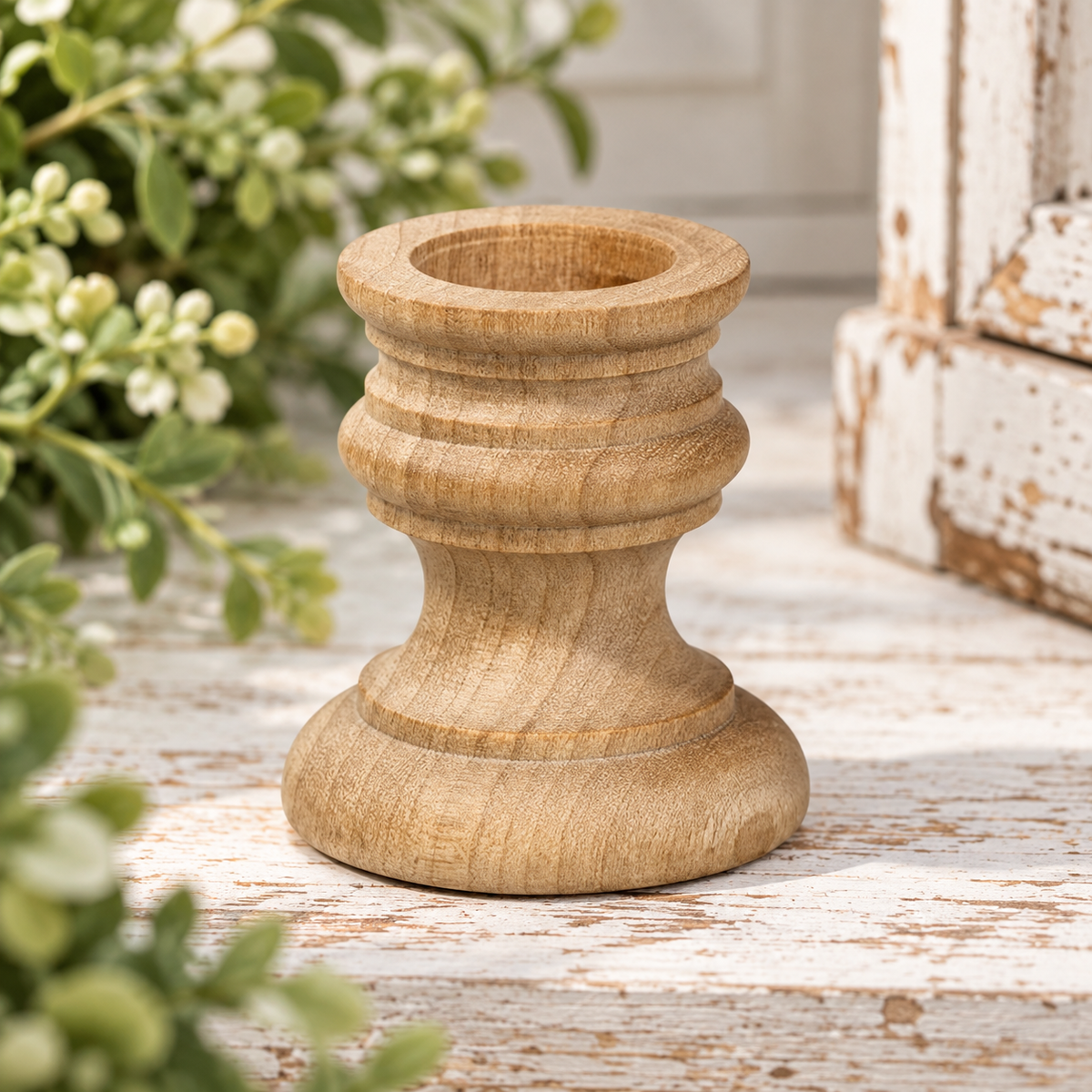 A small carved wooden candle holder or candle stem base with a natural light brown wood finish, set on a distressed white wooden surface with green foliage in the foreground and a rustic white wooden box in the background.