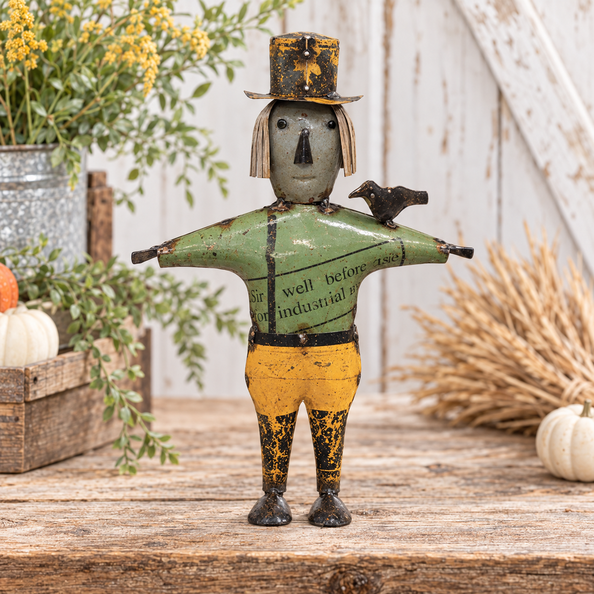 A rustic metal scarecrow figurine with a weathered green shirt, yellow pants, and a black top hat, standing on a wooden surface with autumn-themed props like pumpkins, wheat, and a galvanized bucket in the background.