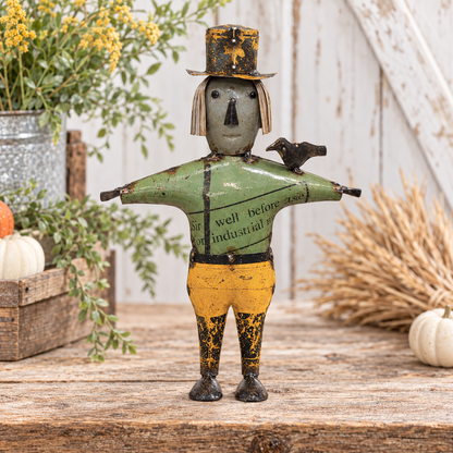 A rustic metal scarecrow figurine with a weathered green shirt, yellow pants, and a black top hat, standing on a wooden surface with autumn-themed props like pumpkins, wheat, and a galvanized bucket in the background.