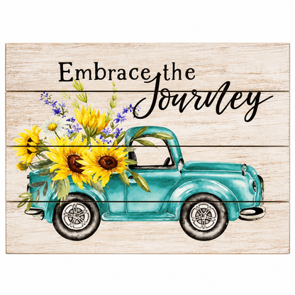 A square decorative sign with a whitewashed wood plank background, featuring a turquoise pickup truck carrying sunflowers and purple wildflowers, with the phrase 'Embrace the Journey' in black script and serif lettering.