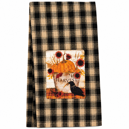 A folded kitchen towel with a black and beige plaid pattern, featuring a central autumn-themed patch with a pumpkin, sunflowers, leaves, and a crow above the word 'HARVEST'.