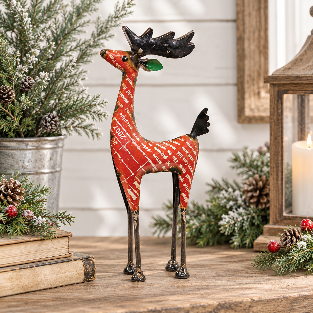 A decorative metal reindeer figurine with a red and white patterned body, black antlers, and a green leaf on its ear, displayed on a rustic wooden table with holiday greenery, books, and a lantern in the background.