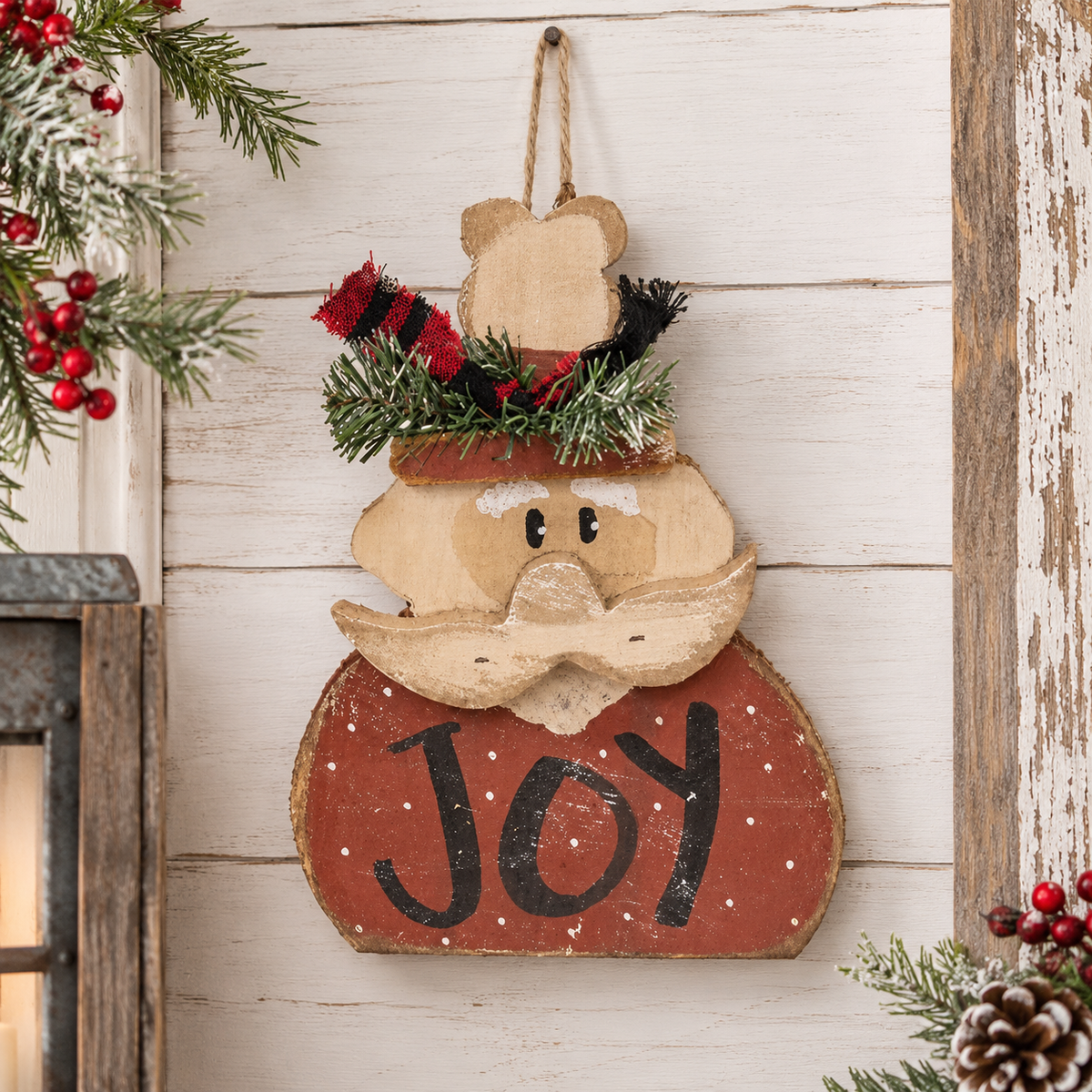 A rustic wooden hanging ornament shaped like a smiling face with a large mustache and the word "JOY" painted on the lower half. The ornament is styled with a red and black striped hat, green pine garland, and white faux snow, hanging against a distressed white wood plank wall with holiday greenery and red berries around it.