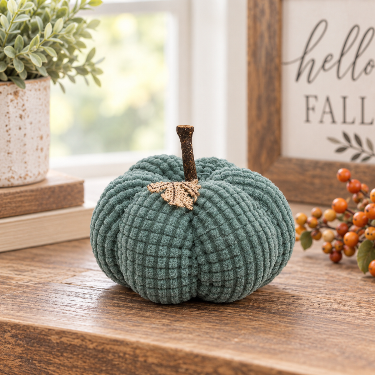 A ribbed fabric decorative pumpkin in muted teal green sits on a wooden table, styled with a potted plant, books, and a fall-themed sign in the background.